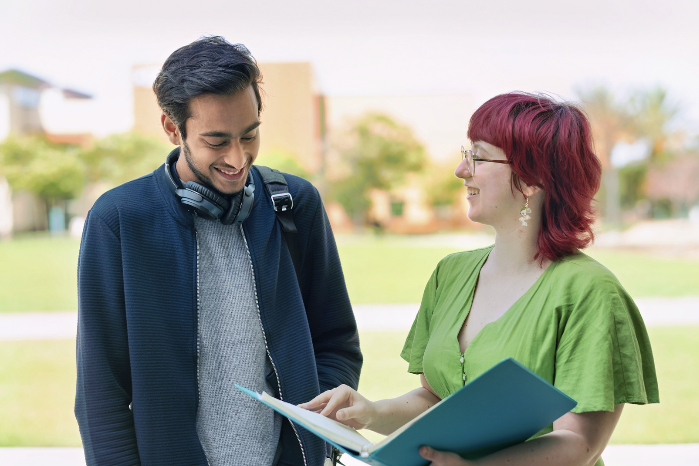 Two students standing outside on a campus, smiling while looking at an open folder one of them is holding.