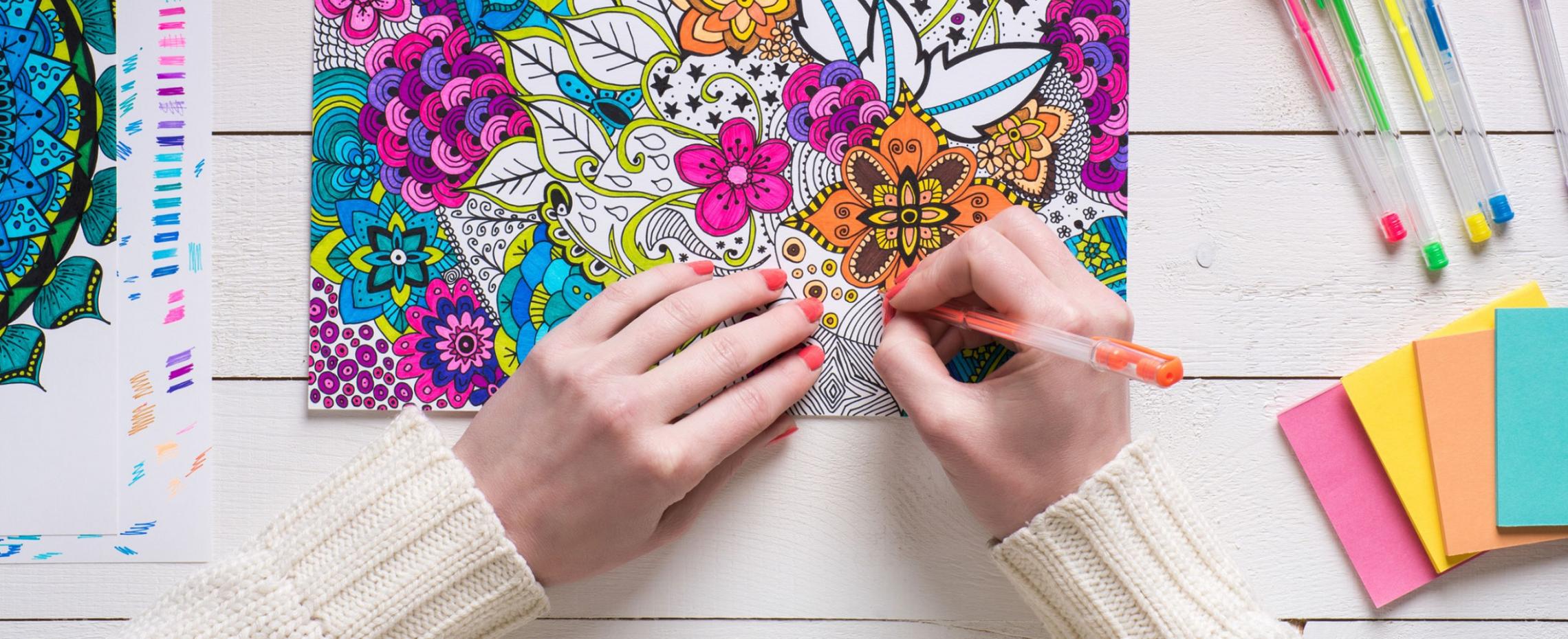A decorative image of hands coloring a mandala coloring page. The image includes small stacks of paper and coloring pens.